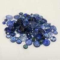 Collection of small loose genuine sapphire gemstones in varied sizes and shapes, showing a range of blue tones from light to dark, arranged on a white background.