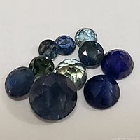 Multiple loose genuine sapphire gemstones in various shades of blue, round and cabochon cuts, arranged on a white surface.
