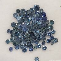 Close-up image showing a pile of small, round faceted blue sapphire gemstones scattered on a white background, displaying various shades of blue and good clarity.