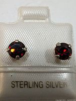 Front view of sterling silver earrings each with a round garnet gemstone in a four-prong setting on a white holder labeled 'STERLING SILVER'