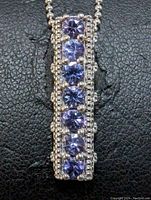 Close-up front view of rectangular pendant with six round-cut tanzanite stones set vertically in sterling silver with beaded borders on black background.