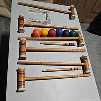 Four wooden mallets arranged front to back with six colored wooden balls placed on one mallet and ten white metal stakes to the side.