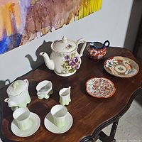 Overall display of tea set items including floral teapot with violet pansy design, red and white creamer, coordinating saucers, and green ombre porcelain items with cups, creamer, and sugar bowl.