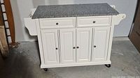 Front view of white kitchen cabinet with polished granite top, two drawers, closed cabinet doors, and caster wheels on the floor.