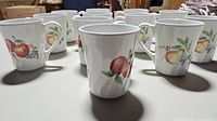 Set of twelve white ceramic mugs with fruit designs, shown in rows on a table