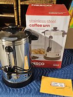 Photo showing Nesco stainless steel coffee urn with black knob lid, spigot, power cord wrapped around base, placed in front of original Nesco box on a blue padded surface.