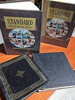 Four stamp albums displayed on wooden surface: two volumes of Standard World Stamp Albums with slipcase, one black album with gold decorative border, one plain black stamp album.