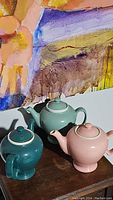 Overview of three vintage ceramic tea kettles in pastel shades: pink, teal, and seafoam green with white trim, displayed on a wooden surface with an artistic background.