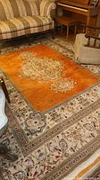 Full view of the handwoven Persian rug placed in a living room with furniture around it, showing the overall condition and color. Orange central field with floral medallions and detailed borders.