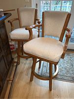 Two wood framed bar stools with upholstered seat and back, positioned side by side near a wooden cabinet and window.
