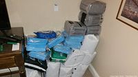 Overview of multiple packages of Depends adult diapers in grey packaging, blue disposable bed pads, underpads, and a green package of wet wipes all stacked near a corner with a wooden table visible.