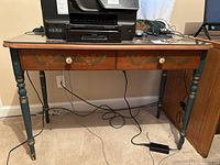 Full frontal view of desk with glass top protector, showing brown wood finish, teal turned legs, two drawers and overall structure