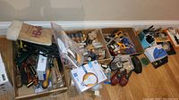 Wide view of multiple hardware items including scissors, screwdrivers, batteries, magnifying glass, two-way radios, and portable fan in box packed loosely in cardboard boxes on floor.
