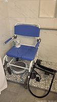 Bariatric shower chair with blue padded seat and backrest, armrests, wheels, and a bucket underneath, shown in a tiled bathroom setting.