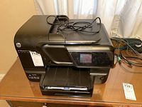 Front and top view of the black HP Officejet Pro 8600 printer showing paper tray, control panel, and cables on device.