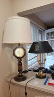 View showing both table lamps on a countertop, one lamp includes a clock base with a cream cloth lampshade, the other has a black shade with a Duke emblem and brass candle holder style base.