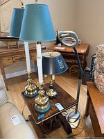 Wide view showing two brass candlestick table lamps with teal shades, black Duke candelabra lamp, and silver magnifier floor lamp