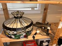 Shelf view showing hanging stained glass lamp with floral and geometric colored glass design next to three flying geese plaque on driftwood base.