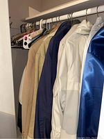View of various men’s outerwear including a beige suit jacket, a blue varsity jacket, and windbreaker jackets hanging on a closet rod.