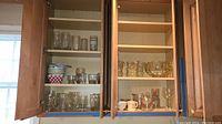 Open wooden cabinet with various glass tumblers, punch bowl set, ceramic mugs, and vintage measuring cups arranged on shelves.