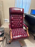 Front view of the burgundy leather office chair showing button tufting, wooden armrests, and wooden base with wheels. Chair is large with high back.