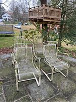 Four wrought iron chairs arranged on outdoor patio, showing mesh seats, scrollwork on backrests, and overall weathered condition with moss and paint chipping.