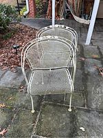 Two white wrought iron outdoor chairs placed on stone pavement outdoors, showing mesh design and slightly weathered paint with chipping and moss.