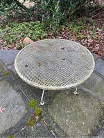 Photo showing entire round wrought iron coffee table with mesh top placed outdoors on stone surface surrounded by garden foliage and a tipped terra cotta pot nearby.