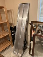Two silver aluminum wheelchair ramps leaning against a wall near a wooden shelf and mirrors. Ramps appear heavy-duty with multiple reinforcements.