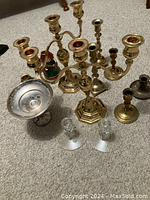 Multiple brass candle operas and candlestick holders arranged on carpeted floor including glass candle holders and silver-tone dish.