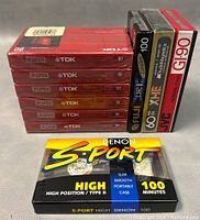 Six sealed TDK 90-minute cassette tapes stacked horizontally with spines labeled, plus three vertically standing tapes from Fuji, TDK, and JVC, and one Denon S-PORT 100-minute tape placed in front.