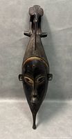 Front view of dark wood carved mask showing bird figure on top and detailed facial features with pigment highlights.
