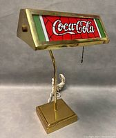 Front angled view of vintage Coca-Cola brass table lamp showing stained glass shade, brass frame with small dents, and attached power cord.
