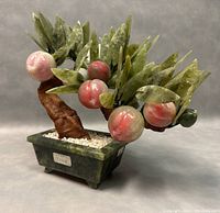Front angled view of vintage jade bonsai peach tree showing pink jade peaches and green jade leaves on textured brown trunk in green jade pot with white pebbles.