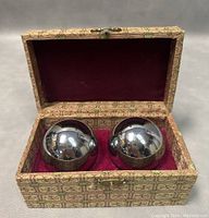 Photo of two stainless steel Baoding balls resting in a red velvet-lined decorative box covered in patterned fabric.