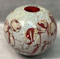 Close-up photo showing the sphere-shaped vase with reversed painted abstract designs in red and beige on a gray-white textured background. The circular top opening reveals the red interior. Minor surface wear visible.