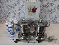 Photo of two Home & Gift stainless steel fondue sets each with glass lids and bases, plus five fuel bottles and multiple fondue forks on a countertop.