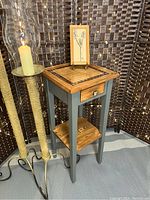 The accent table shown fully assembled with a decorative framed art and candle on top, in front of a woven screen.