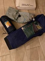 All three items folded and packed in their bags on a tiled floor: one blue bag with sleeping bag label, one navy blue bag for folding bed frame, and one gray air mattress.