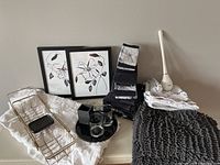 Photo showing part of the bathroom starter kit including two black and white framed prints, stacked towels in black, white and gray, a white toilet brush, bath tray with black soap dish, two black glasses and a toothbrush holder on a black tray, and gray mats.