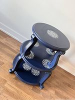 Navy blue three-tier circular accent table with cream decorative motifs on each tier, viewed from an angle.