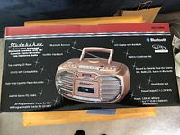 Box back with product image and feature list including Bluetooth receiver, top loading CD player, cassette recorder, AM/FM radio, LCD display with backlight, headphone and aux inputs.