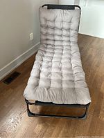 Full view of the reclining camping cot with tufted removable mattress and pillow in light grey color on a black metal frame on wooden floor, showing the angled reclining position.