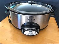 Front view of the Rival Crock-Pot showing stainless steel housing, black trim, knob dial control panel and glass lid with handle.