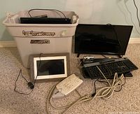 Overall lot with electronics including plastic bin filled with electronic devices, TV, keyboard, camera, digital photo frame, and power strip on carpet floor.