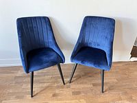 Two royal blue velvet chairs with vertical channel tufting on back and black legs shown side by side on hardwood floor against white wall.