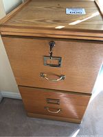 Front view of the wooden filing cabinet showing two drawers with brass handles and a key in the lock of the top drawer.