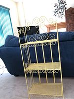 Front view of the yellow metal shelving unit showing three wire shelves and intricate scrollwork on the top and sides, with a blue couch in the background.