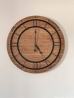 Front view of large wooden wall clock mounted on wall showing wood grain and black metal Roman numerals with clock hands at 5 o'clock.