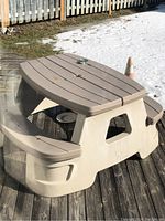 Full view of the beige plastic adult patio table with attached benches on a wooden deck, showing its shape and design.